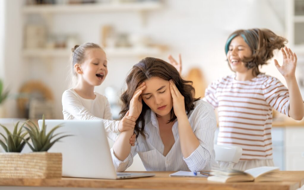 Madre estresada intentando trabajar en la computadora mientras sus hijos la interrumpen constantemente.